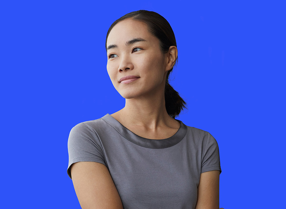 Asian woman on a solid blue background wearing a gray t-shirt and a ponytail looking off into the distance